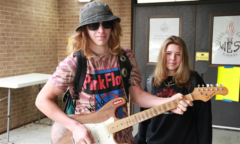 A student holding a guitar stands next to another student